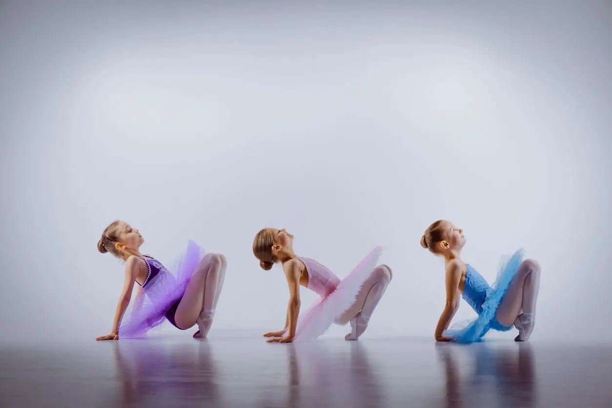 Three little ballet girls sitting  in multicolored tutus and pointe shoes together on white background